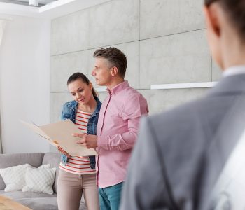 A man and a woman holding an open folder and reviewing paperwork, while standing in a room next to a real estate agent.