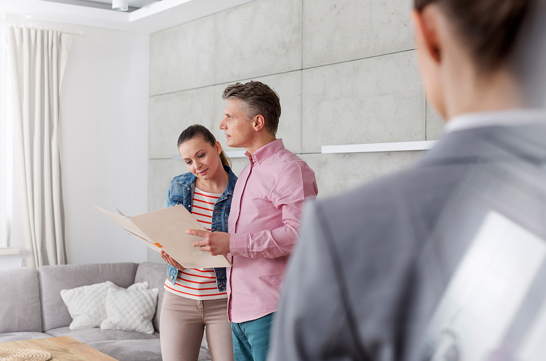 A man and a woman holding an open folder and reviewing paperwork, while standing in a room next to a real estate agent.